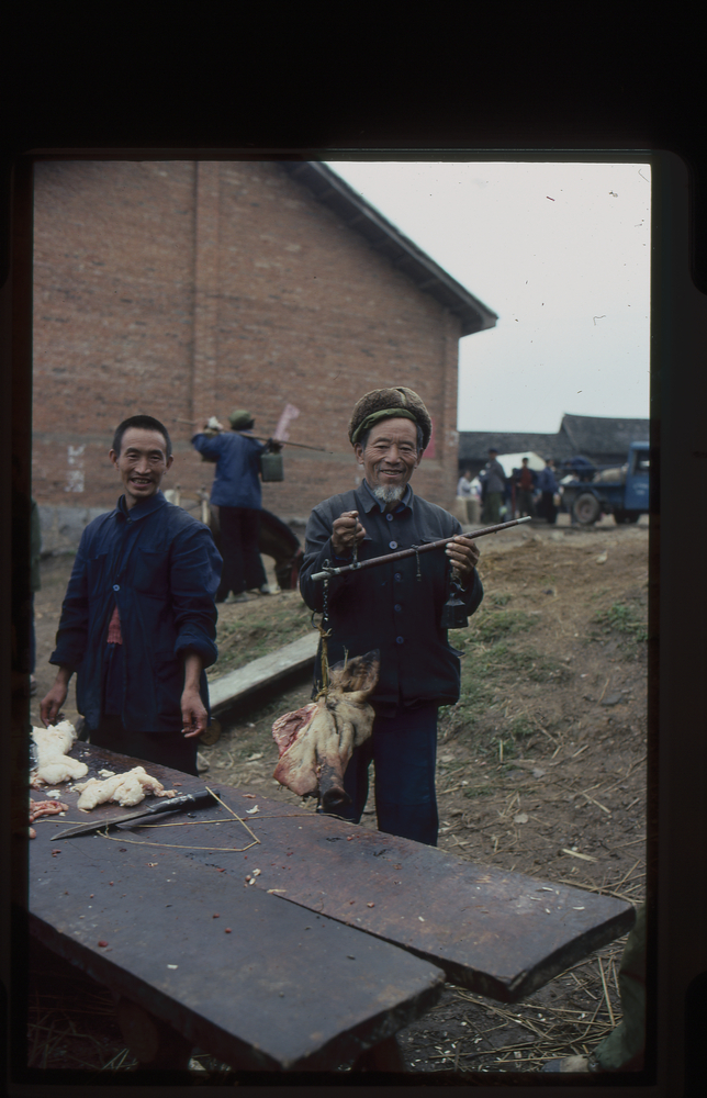 Image of 35mm slide: Friday 28th continued. Butcher weighing meat in the open air