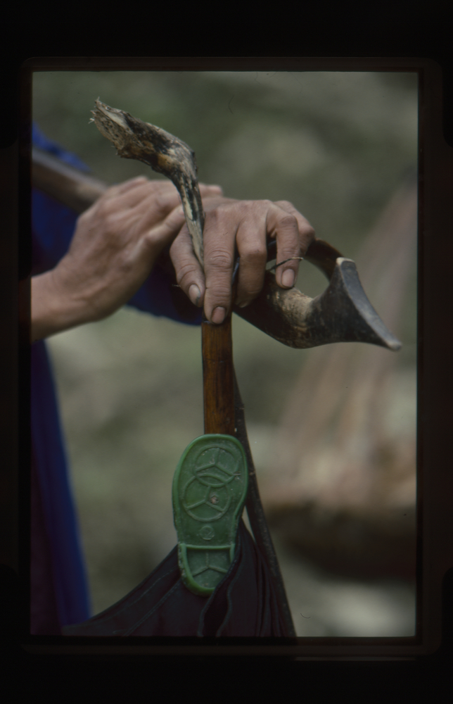 Image of 35mm slide: The same woman holding a walking stick, pole for bag, with plimsoll sticking out of bag.