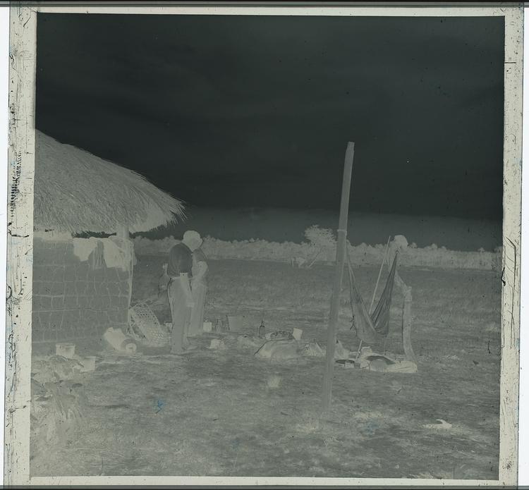 Image of Black and white negative of people outside hut with household objects and hammock, looking out in the middle of a field