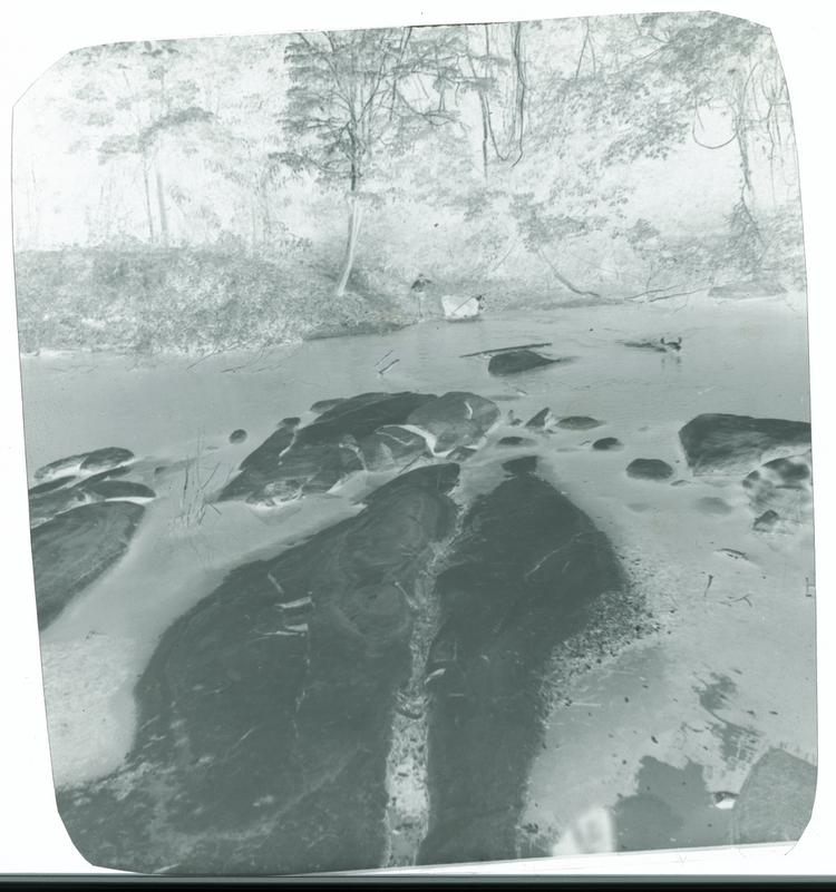 image of Black and white negative of large rocks in river with man herding cattle across water in background