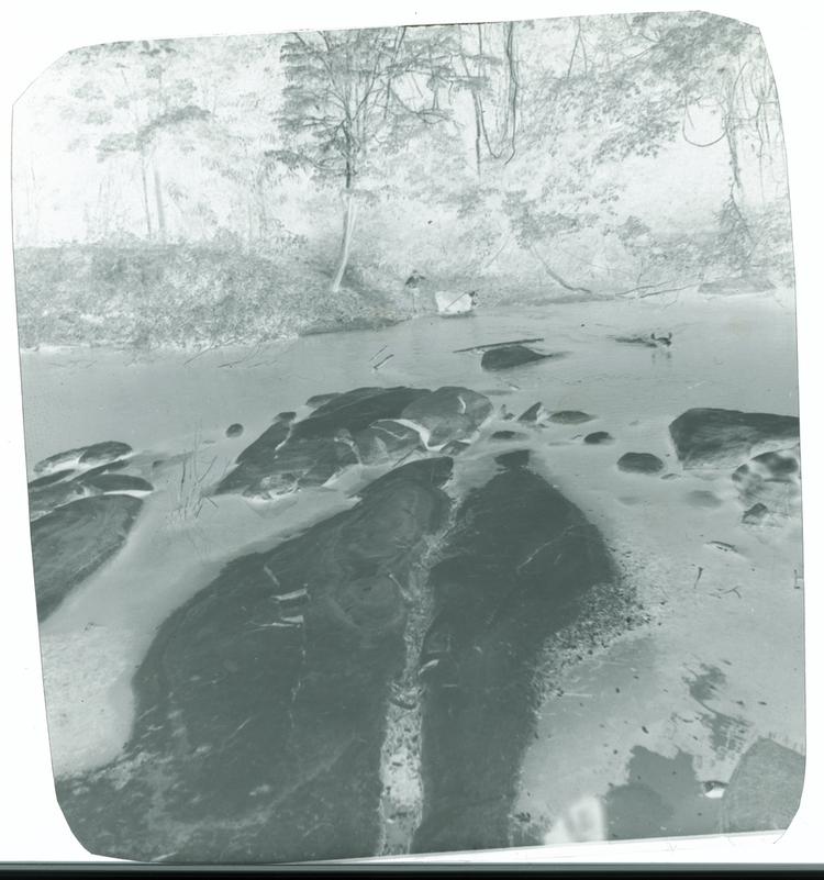 Image of Black and white negative of large rocks in river with man herding cattle across water in background