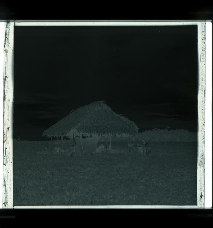 image of Black and white negative of man sitting at table next to large grass hut in open field with many supplies