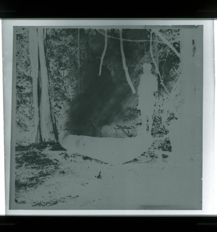 image of Black and white negative of people working on canoe with one man standing on top of canoe in silhouette in middle of dramatically-lit forest