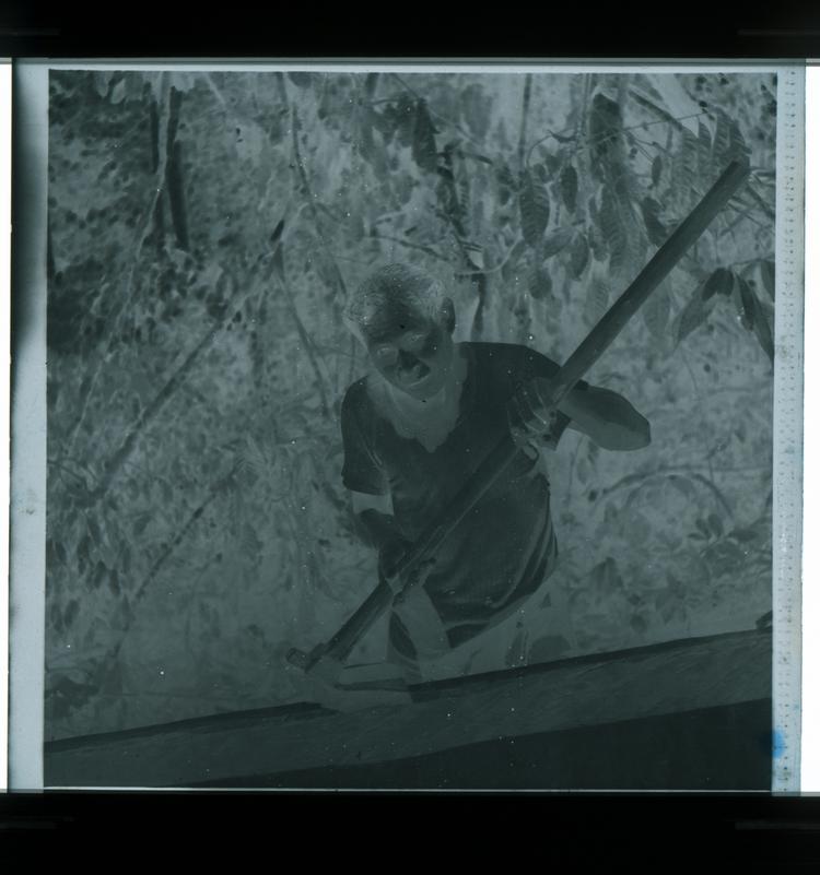 image of Black and white negative of man coyly smiling at camera holding gardening hoe, mid shot