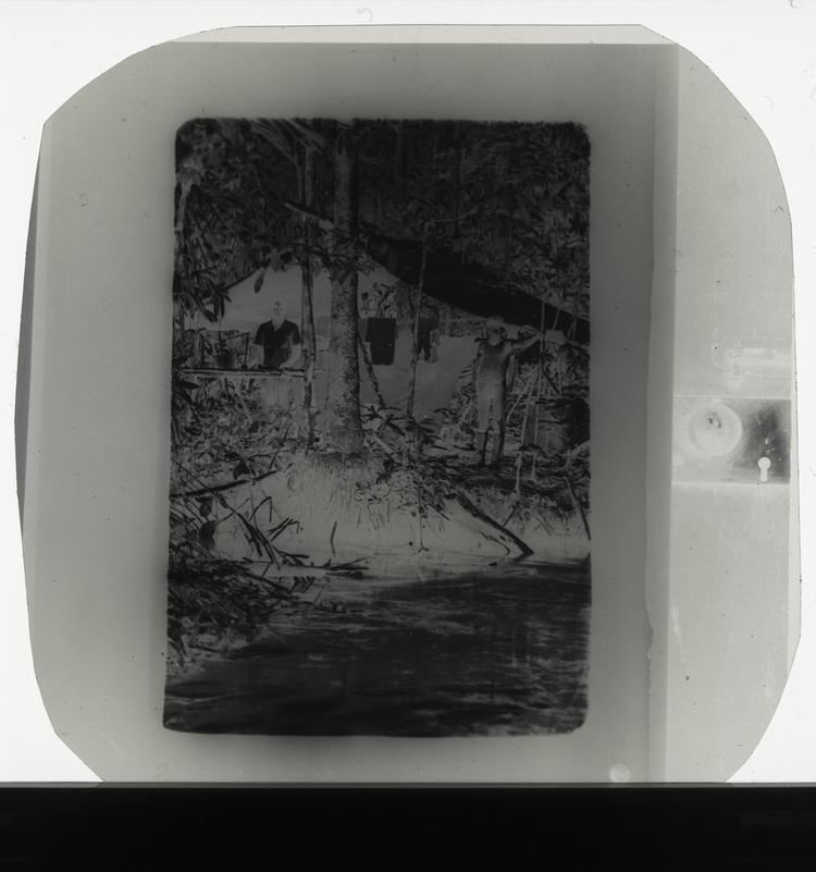 Image of Black and white negative of young Wai Wai tribe member and expedition member standing in front of hut along water preparing food with clothes hanging in background