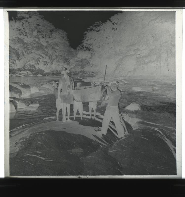 Image of Black and white negative of expedition members carrying boat up onto boulders from tree-lined river