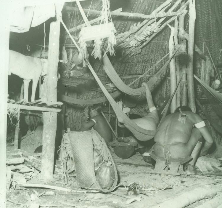 Image of Black and white transparency. Interior with hammocks, three people and a dog. Positive of slide 3. This image is included in Nicholas Guppy's 1958 book and is entitled 'Wai-Wai Interior'