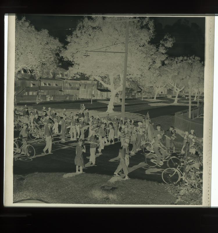 image of Black and white negative of steel drummers and cyclists in parade with standard bearer, far away