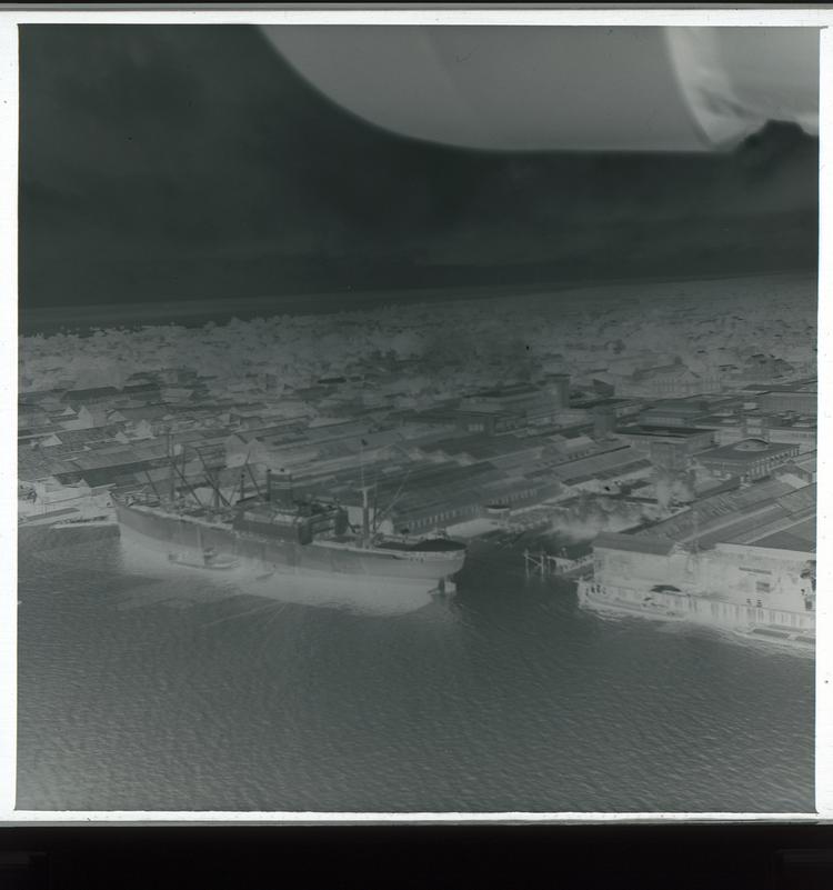 Image of Black and white negative view from air of industrial-looking part of town on coastline with boat in water