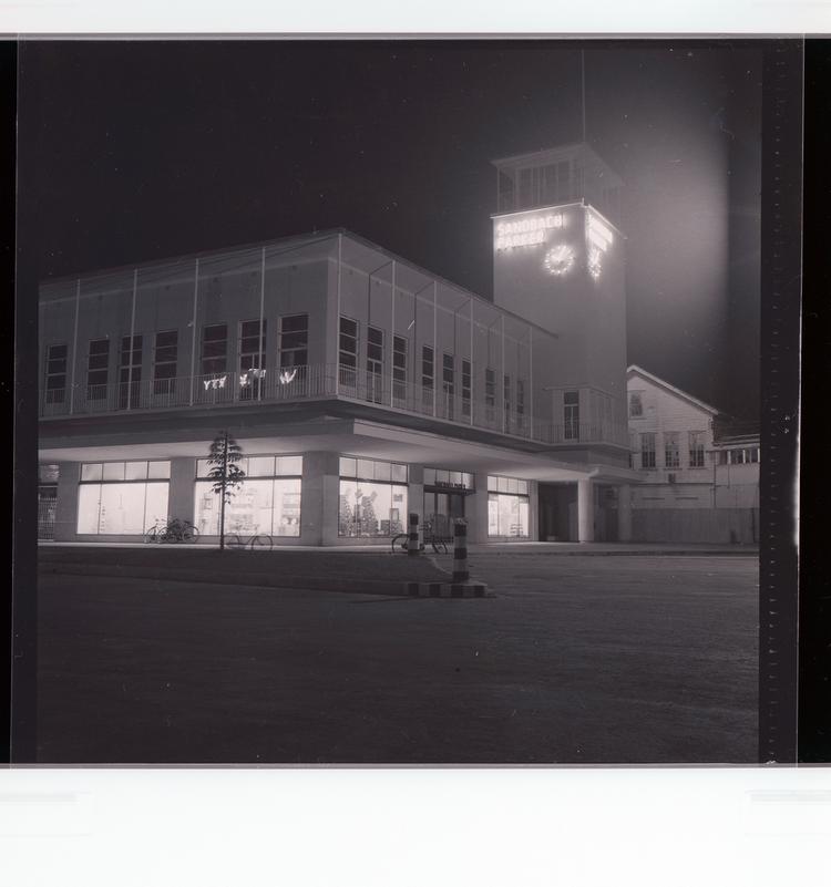 Image of Black and white negative view at night of building with 'Sandback Parker' written on clock tower with bicycles in front