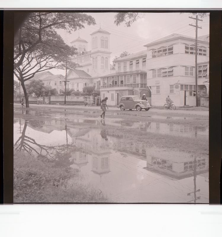 image of Black and white negative view of flooded tree-lined street with buildings, with people walking and cycling