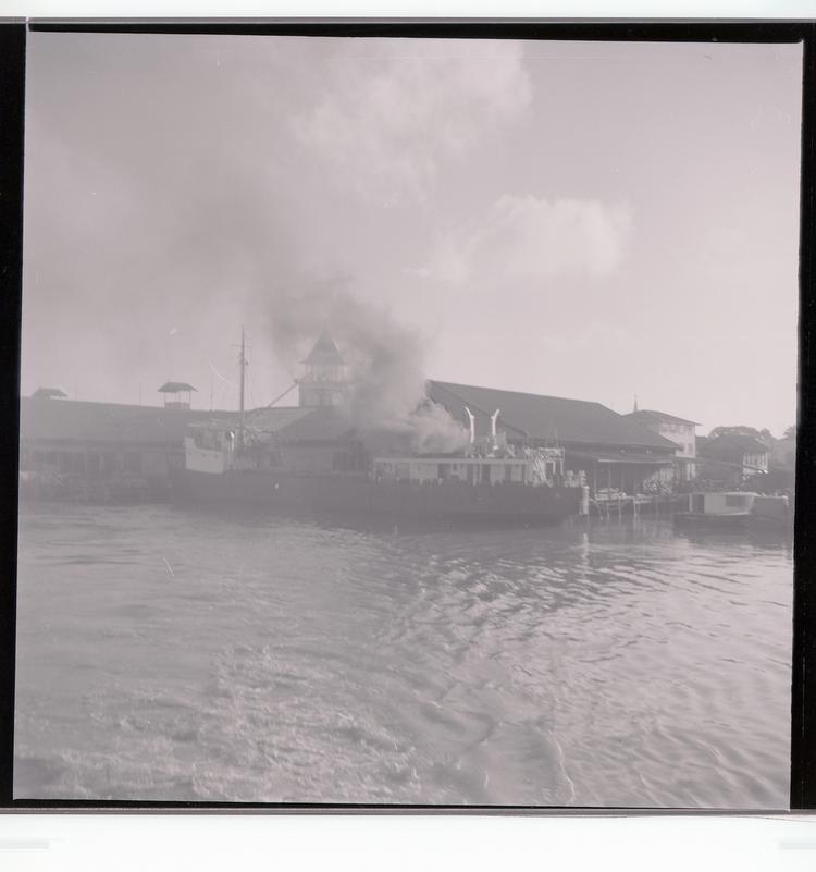 image of Black and white negative of ship (with billowing smoke) docked at port building, view from out in the water