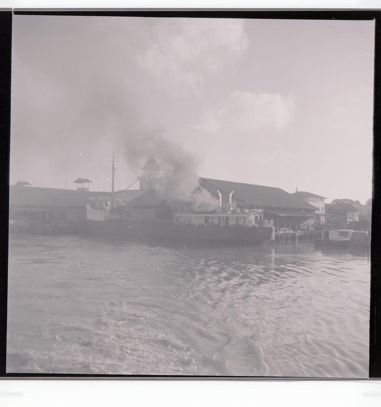 Image of Black and white negative of ship (with billowing smoke) docked at port building, view from out in the water