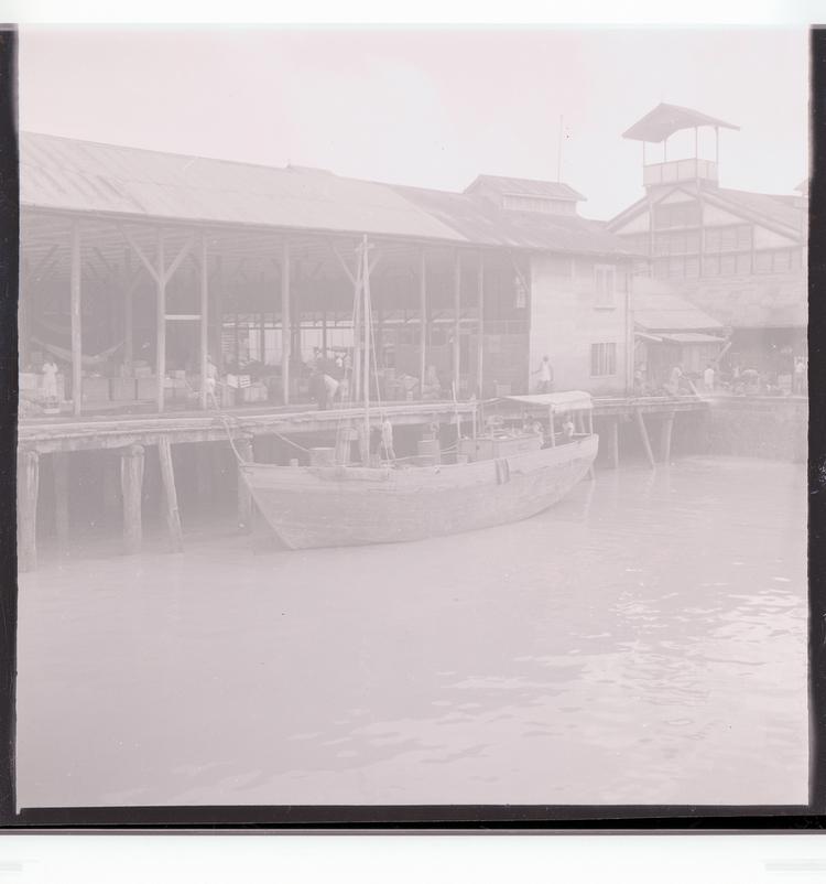 image of Black and white negative of docked boat, loading/unloading goods, port building in background