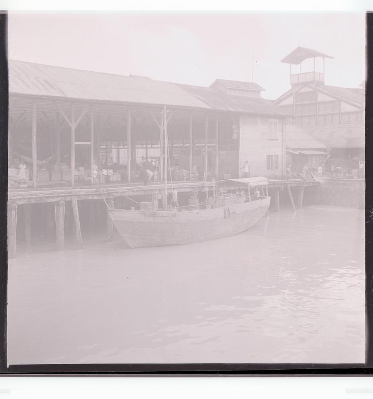 Image of Black and white negative of docked boat, loading/unloading goods, port building in background