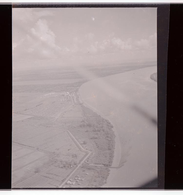 image of Black and white negative of large winding river/coastline, cultivated fields, and buildings from air with part of wing in frame