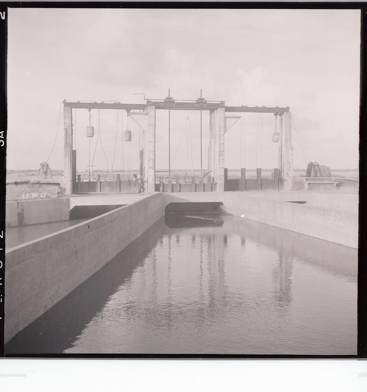 Image of Black and white negative of dam-like system with three sections and bridge/pulleys spanning water with man bending over