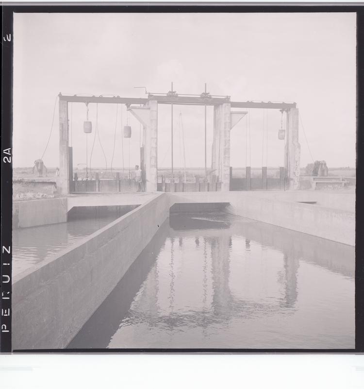 image of Black and white negative of dam-like system with three sections and bridge/pulleys spanning water with man standing upright