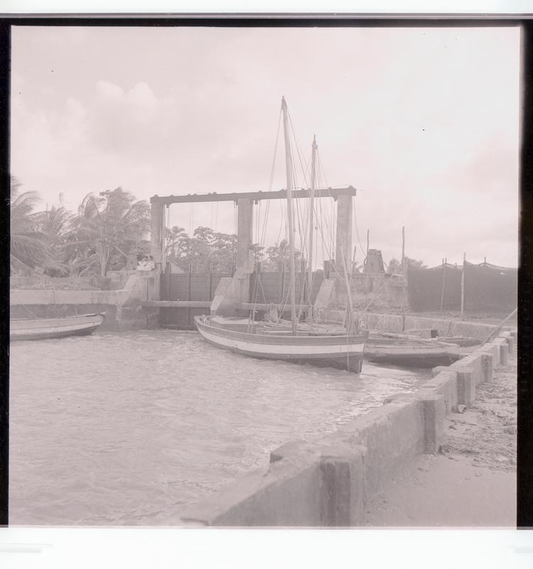 image of Black and white negative of boats moored next to barrier for water/boat passage