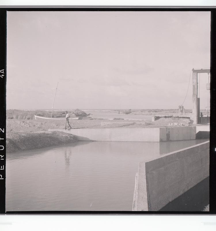 image of Black and white negative of boat and man walking near barrier for water looking toward water