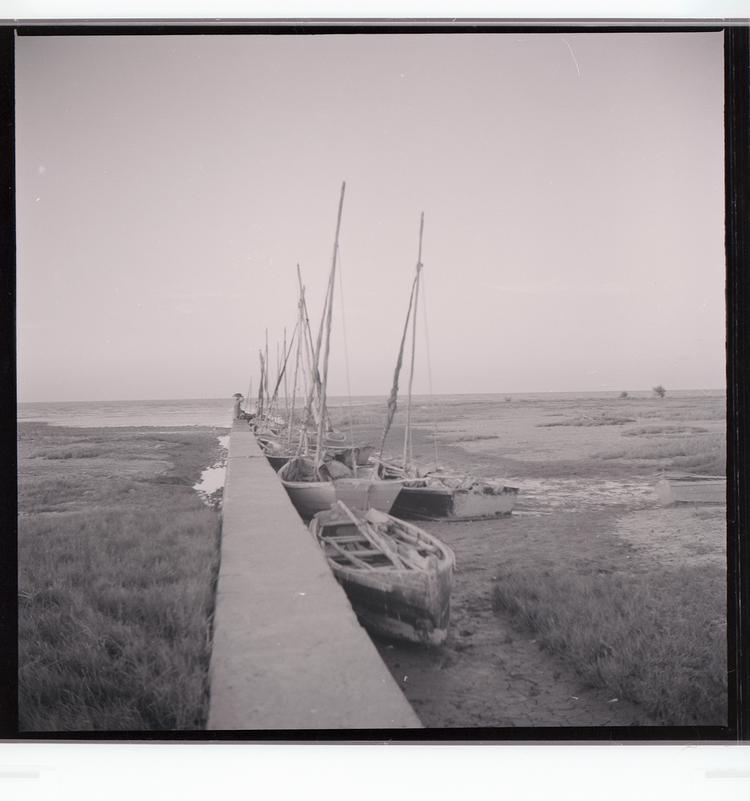image of Black and white negative of boats pulled up on boggy land, tied to wall protruding out into water