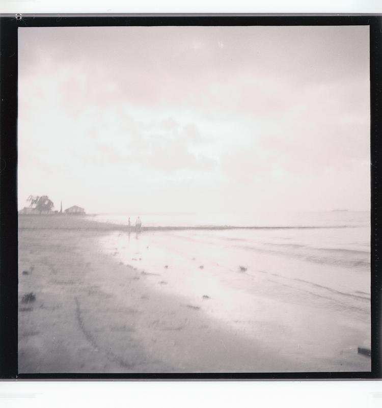 image of Black and white negative of people standing on beach (view from farther down beach), outcropping with building in distance