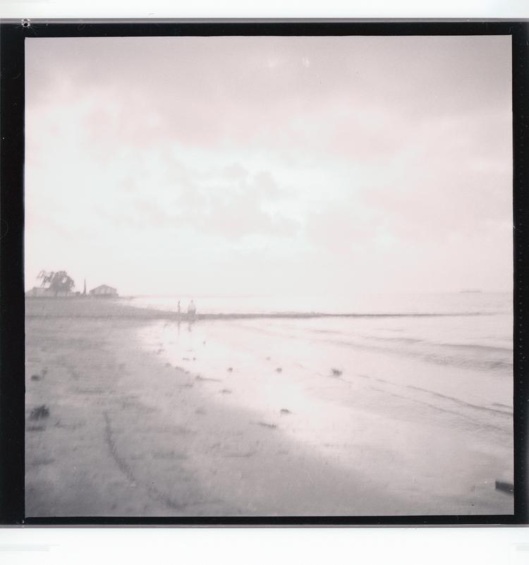 Image of Black and white negative of people standing on beach (view from farther down beach), outcropping with building in distance