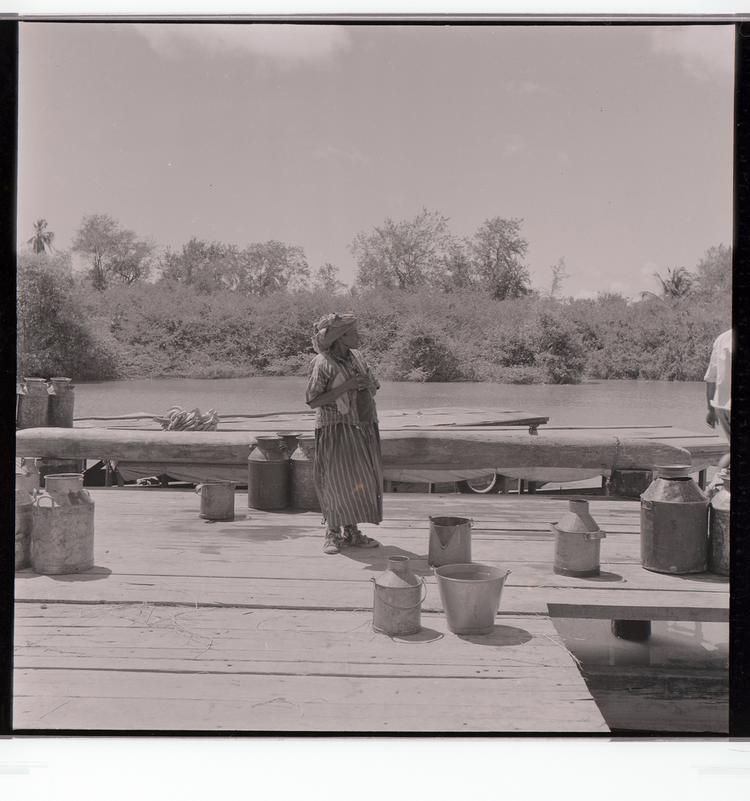 Image of Black and white negative of older woman standing on dock among buckets/containers next to boats docked