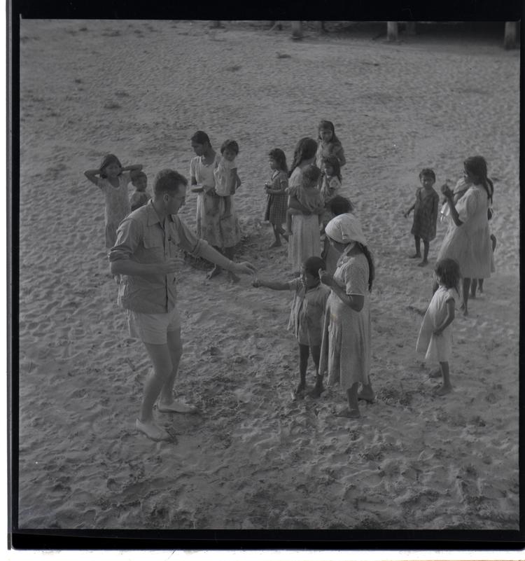 Image of Black and white medium format negative (scanned positive) of adults and children on a sandy area - a man profers medicine to a mother aand child