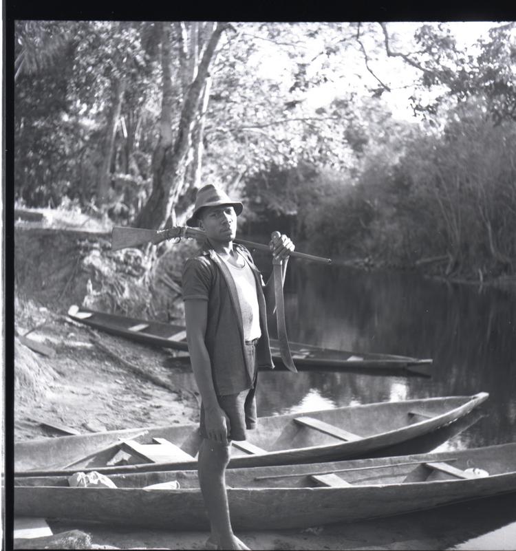 image of Black and white medium format negative (scanned positive) of a man standing by a river with a gun on one shoulder with wooden canoes at his feet