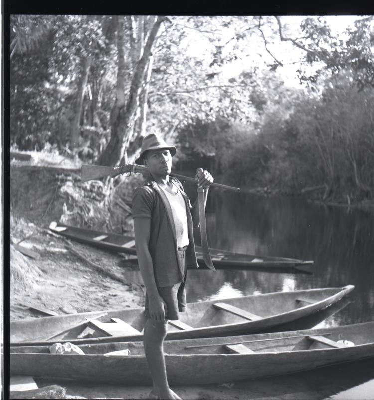 Image of Black and white medium format negative (scanned positive) of a man standing by a river with a gun on one shoulder with wooden canoes at his feet