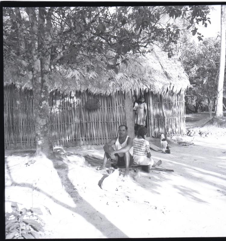 Image of Black and white medium format negative (scanned positive) of a man sitting outside a thatched hut with a woman  (two girls are standing in the hut doorway)