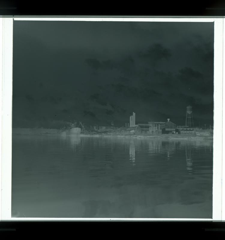 Image of Black and white negative of multiple boats docked on river next to factory (view from far away on water featuring factory)