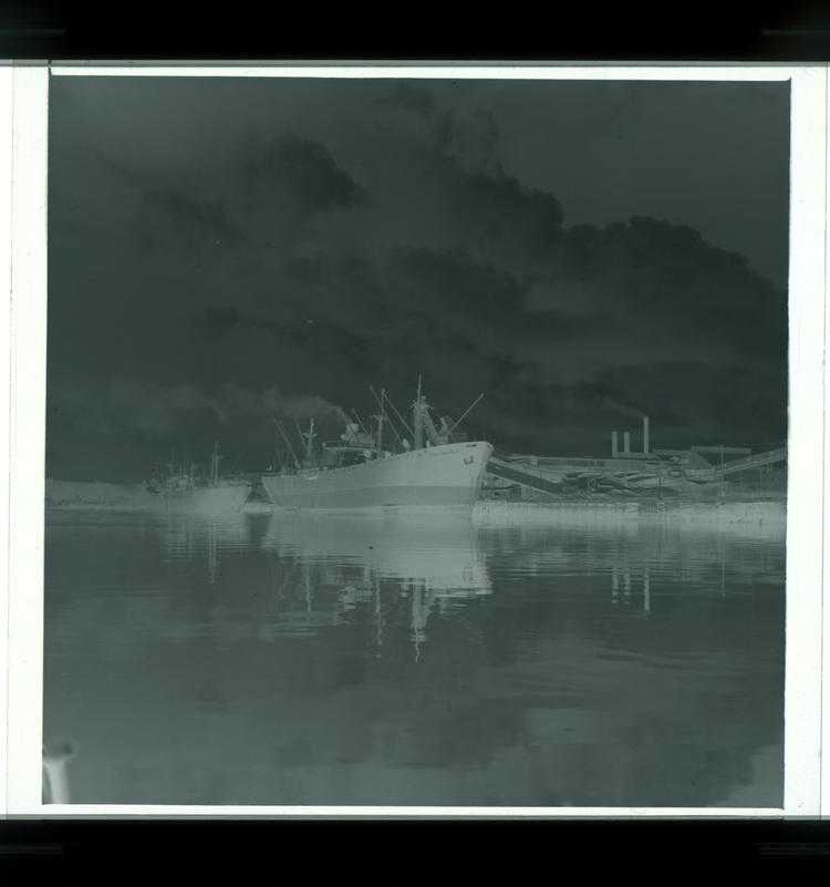 Image of Black and white negative of multiple boats docked on river next to factory (midway view from water featuring both factory and boats)