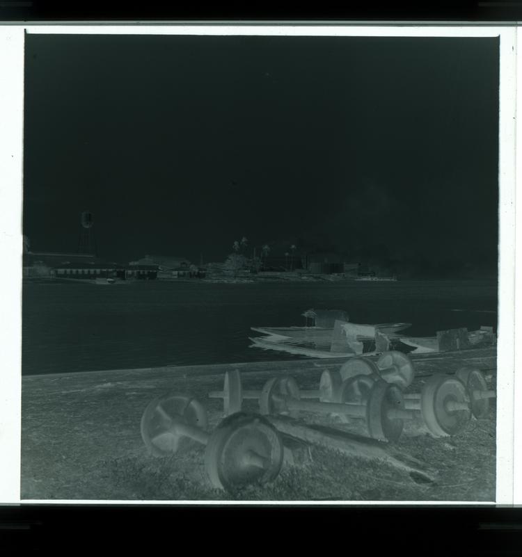 image of Black and white negative of side of river with metal railroad pieces and factory buildings across river in background