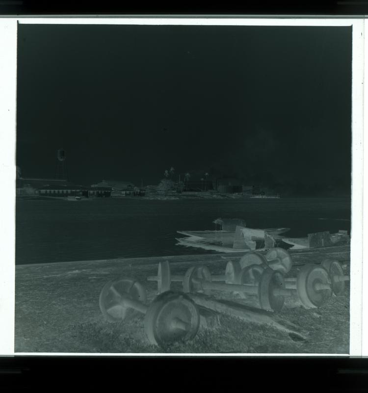 Image of Black and white negative of side of river with metal railroad pieces and factory buildings across river in background