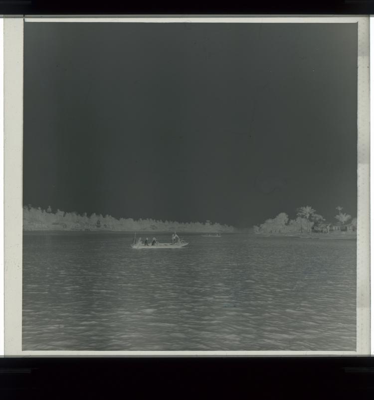 image of Black and white negative of men in large row boat (with one man standing at prow) in middle of river with houses on stilts to the right [far away]