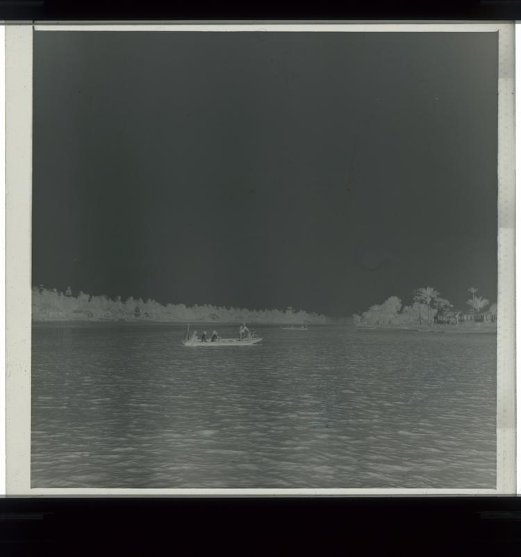 Image of Black and white negative of men in large row boat (with one man standing at prow) in middle of river with houses on stilts to the right [far away]