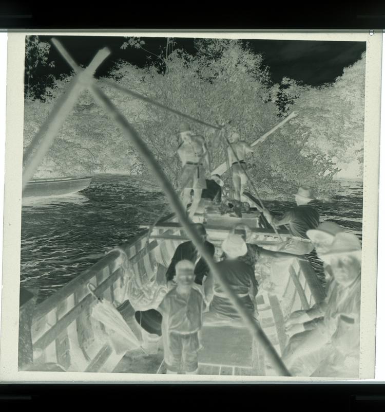 image of Black and white negative of deep boat with a few people in it on river (view from on the boat itself)