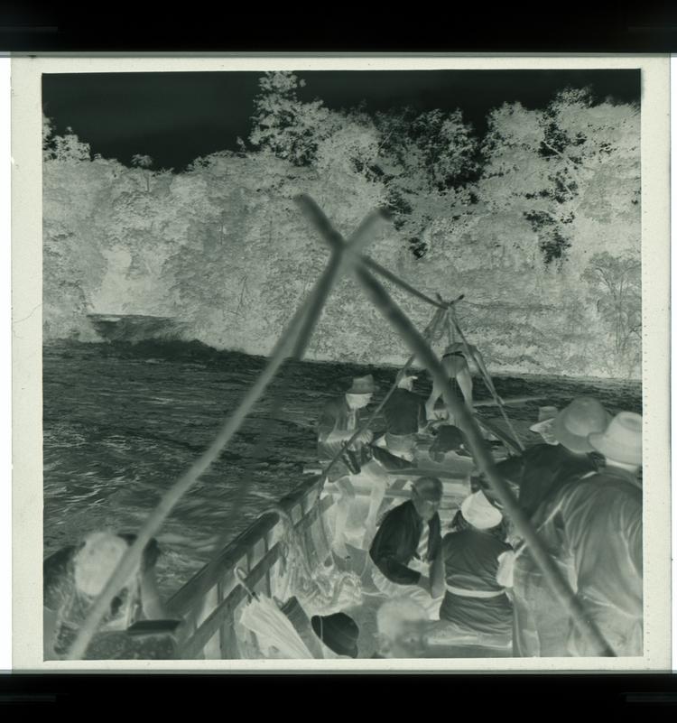 image of Black and white negative of deep boat with a few people in it on river in front of swiftly moving water (view from on the boat itself)