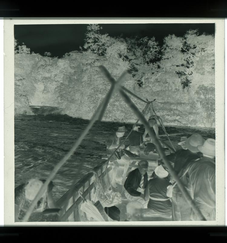 Image of Black and white negative of deep boat with a few people in it on river in front of swiftly moving water (view from on the boat itself)