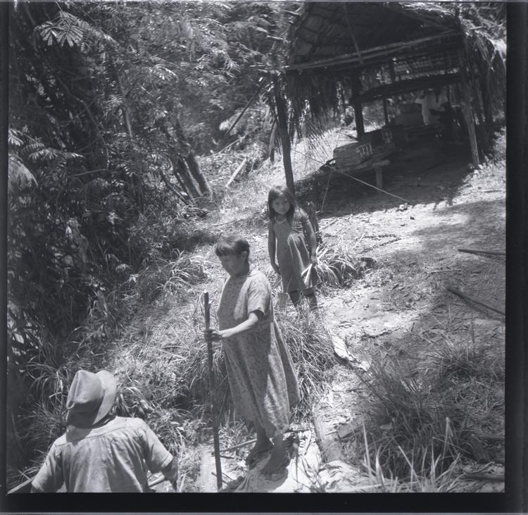 Black and white medium format negative of Indian woman and child with back of a man in a hat in the foreground shelter/building in background