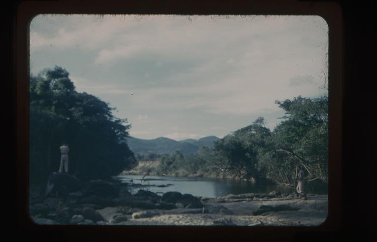 Colour slide of two men by a calm river bordered by trees with mountains in the distance (one man on a rock -perhaps taking a photoraph)
