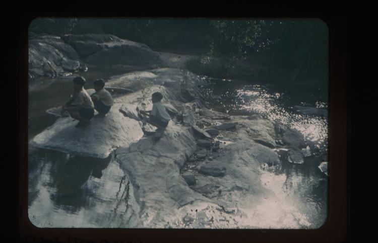 Colour slide of three boys crouching on rocks in a river
