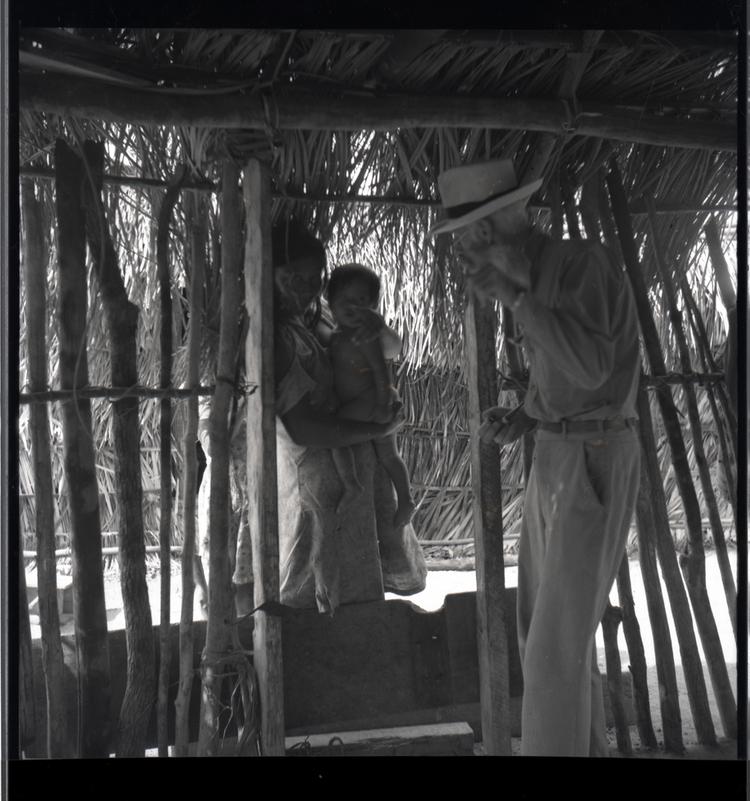 Image of Black and white medium format negative of man talking to a woman holding a child in a wooden hut