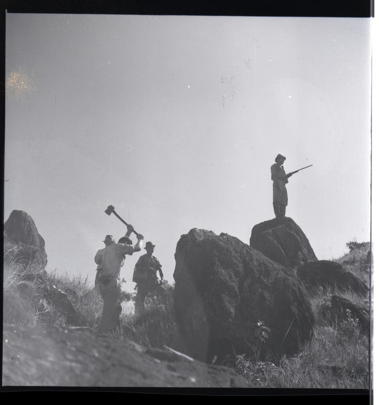 Black and white medium format negative of Group of men breaking stones with one on a boulder possibly holding a gun