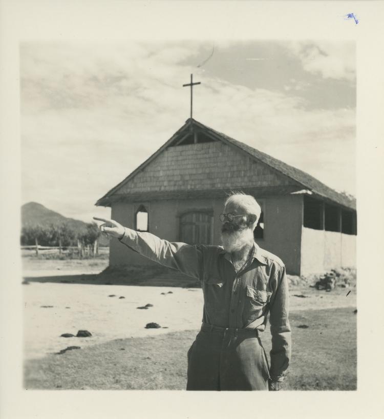 Image of Black and white print of man standing outside simple church building with cross on roof