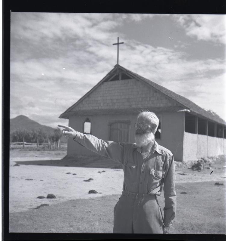 Black and white medium format negative of man standing outside simple church building with cross on roof