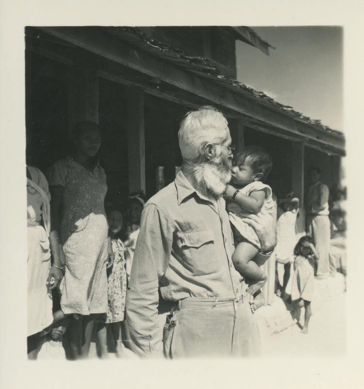 Black and white print of man in front of a building holding a baby (people behind in shade of building)