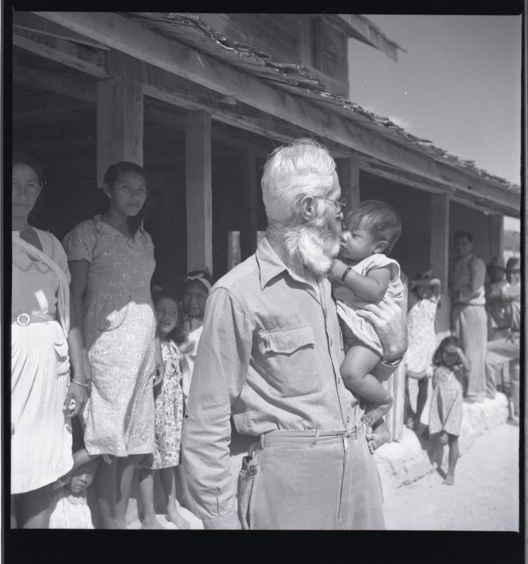 Image of Black and white medium format negative of man in front of a building holding a baby (people behind in shade of building)